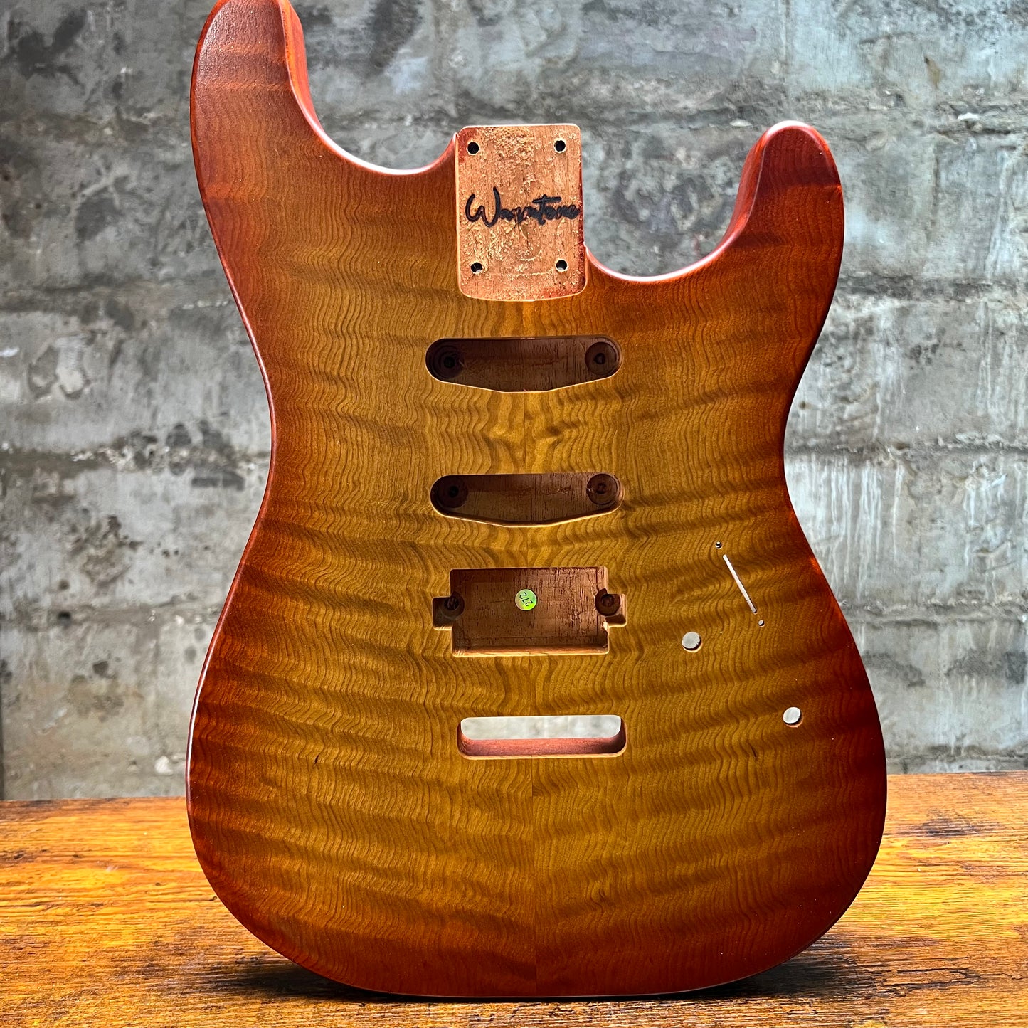A custom electric guitar body made of figured maple wood with a warm honey-brown finish, featuring a hand-written 'Weston' brand patch on the headstock area, set against a rustic gray brick wall and wooden table background.