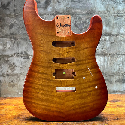 A custom electric guitar body made of figured maple wood with a warm honey-brown finish, featuring a hand-written 'Weston' brand patch on the headstock area, set against a rustic gray brick wall and wooden table background.