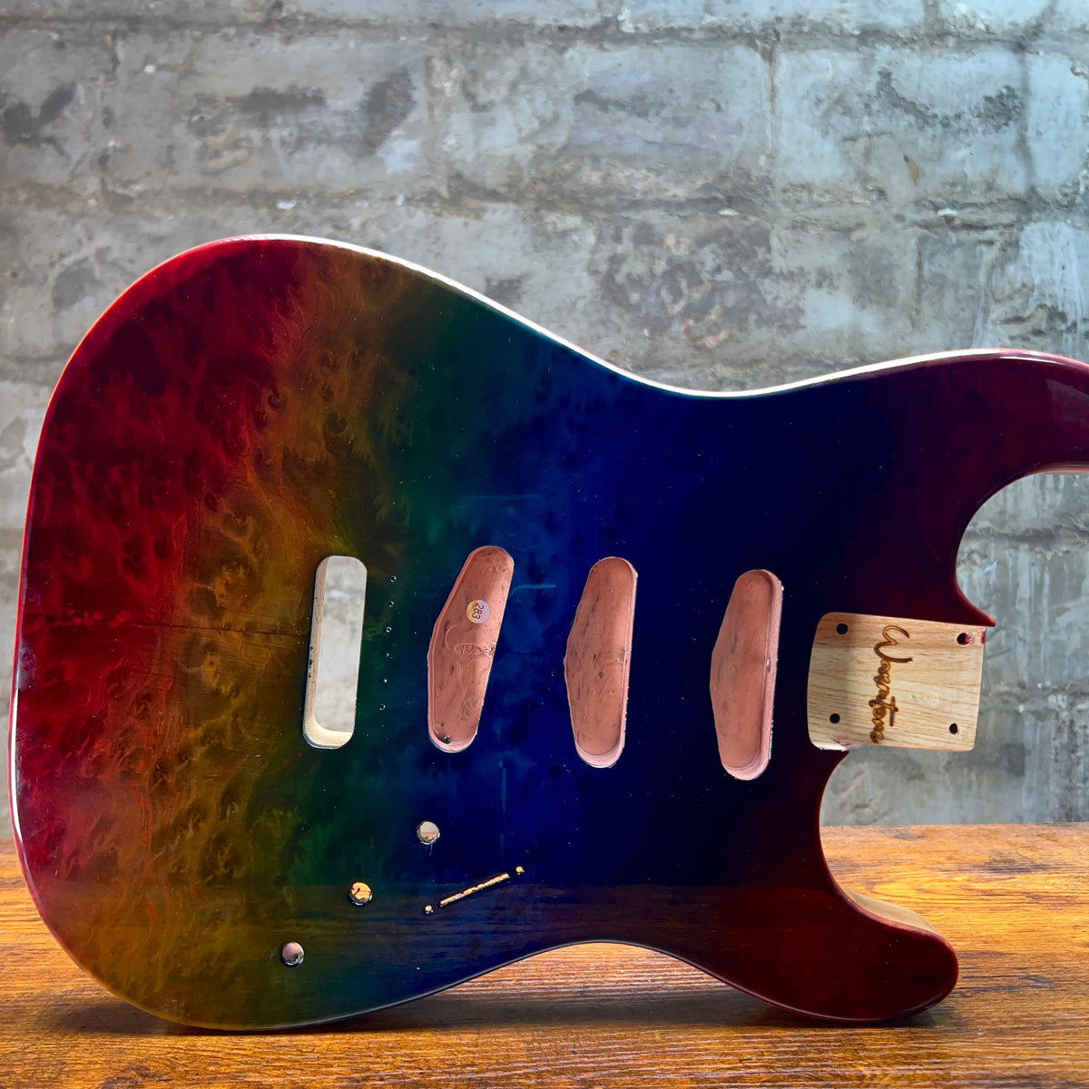 A custom electric guitar body pickguard with a vibrant rainbow gradient finish, resting on a wooden surface against a gray brick wall background.