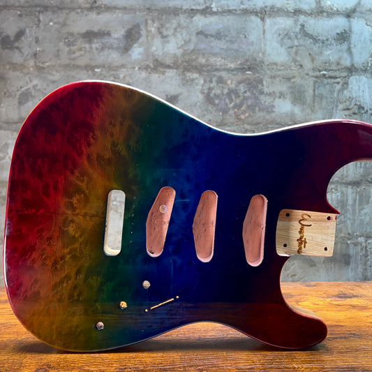 A custom electric guitar body pickguard with a vibrant rainbow gradient finish, resting on a wooden surface against a gray brick wall background.