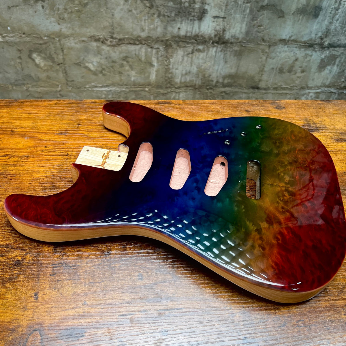 A custom electric guitar body plate with a vibrant rainbow gradient finish, resting on a rustic wooden table against a gray brick wall.