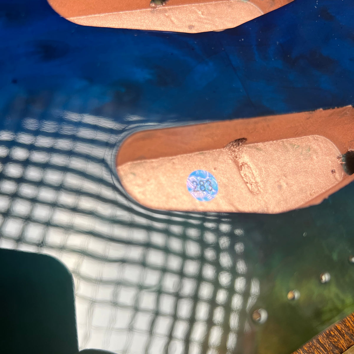 Close-up of a blue electric guitar body showing the wood grain around the pickups, with a small blue circular sticker on one pickup cover.