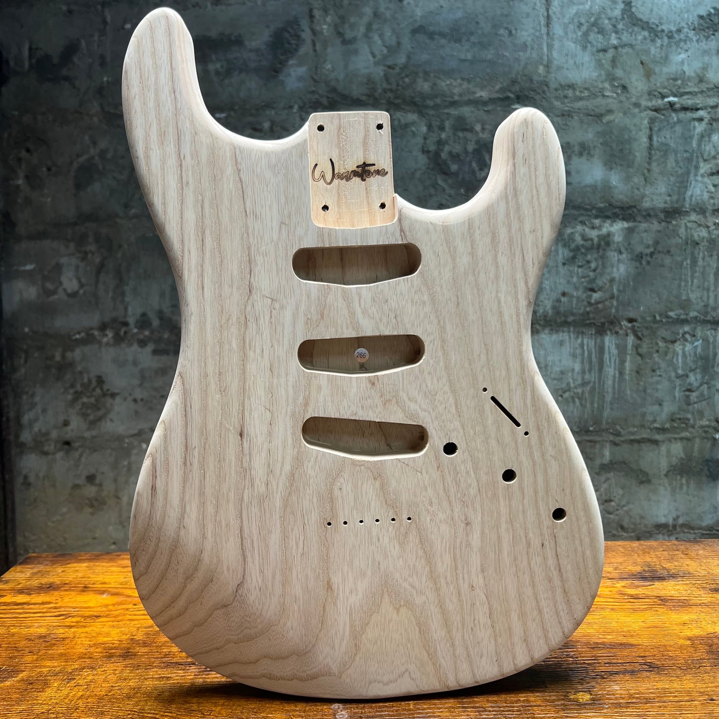 Unfinished swamp ash guitar body on a wooden surface with a textured wall background