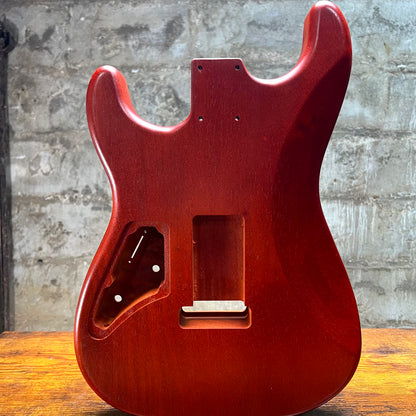 A glossy reddish-brown electric guitar body shell, likely a Strat-style cutaway, standing upright on a wooden surface against a gray concrete wall.