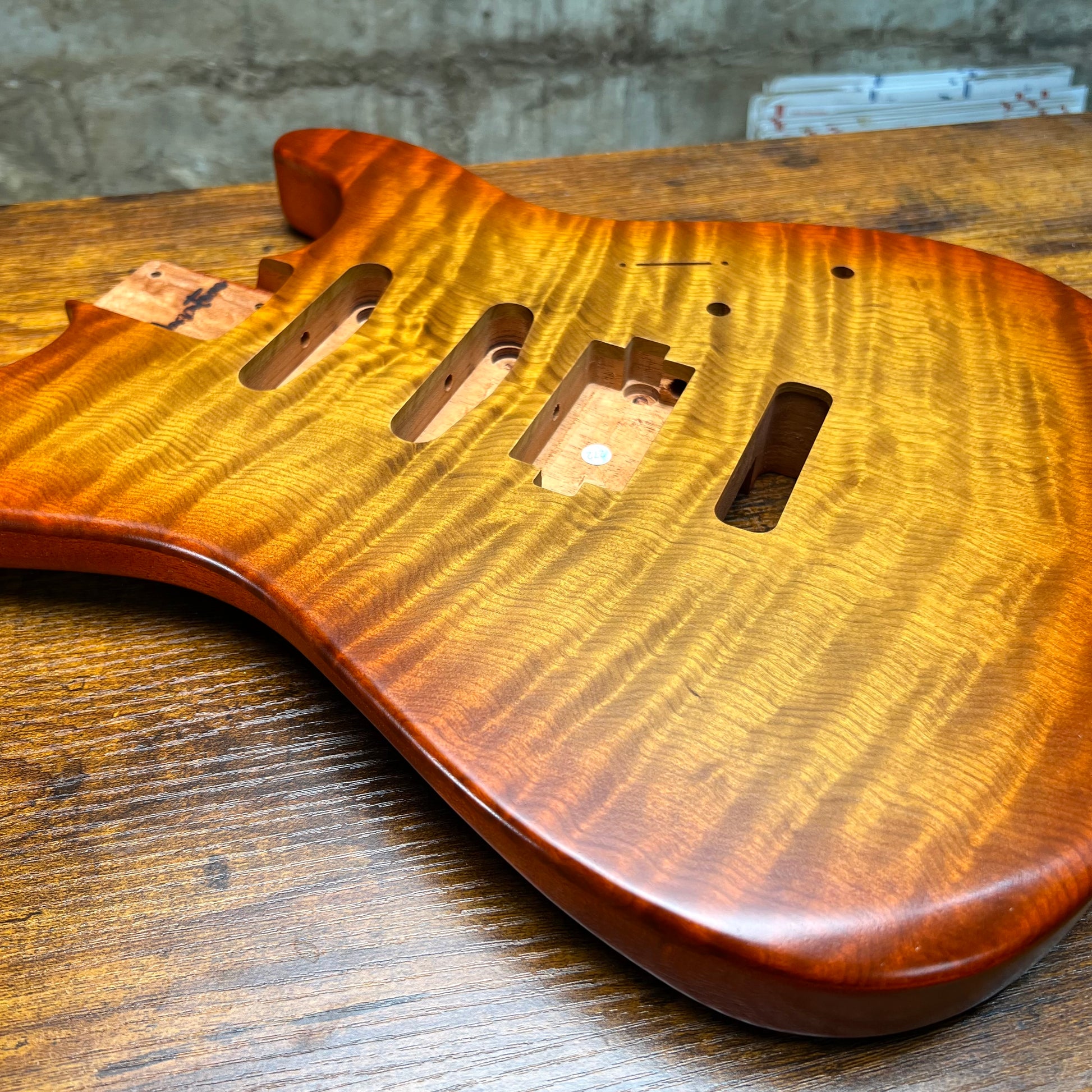 A close-up of a custom electric guitar body made from figured maple wood, showing a glossy natural finish and pre-cut hardware slots, resting on a wooden workbench.
