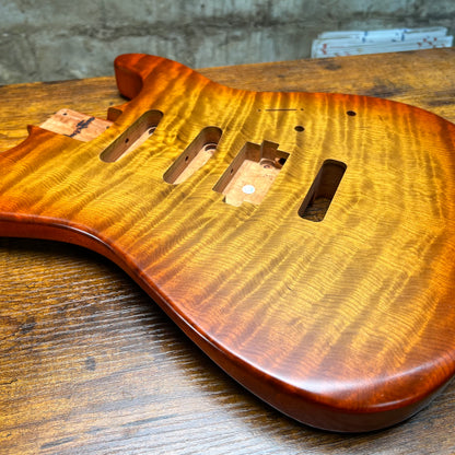 A close-up of a custom electric guitar body made from figured maple wood, showing a glossy natural finish and pre-cut hardware slots, resting on a wooden workbench.